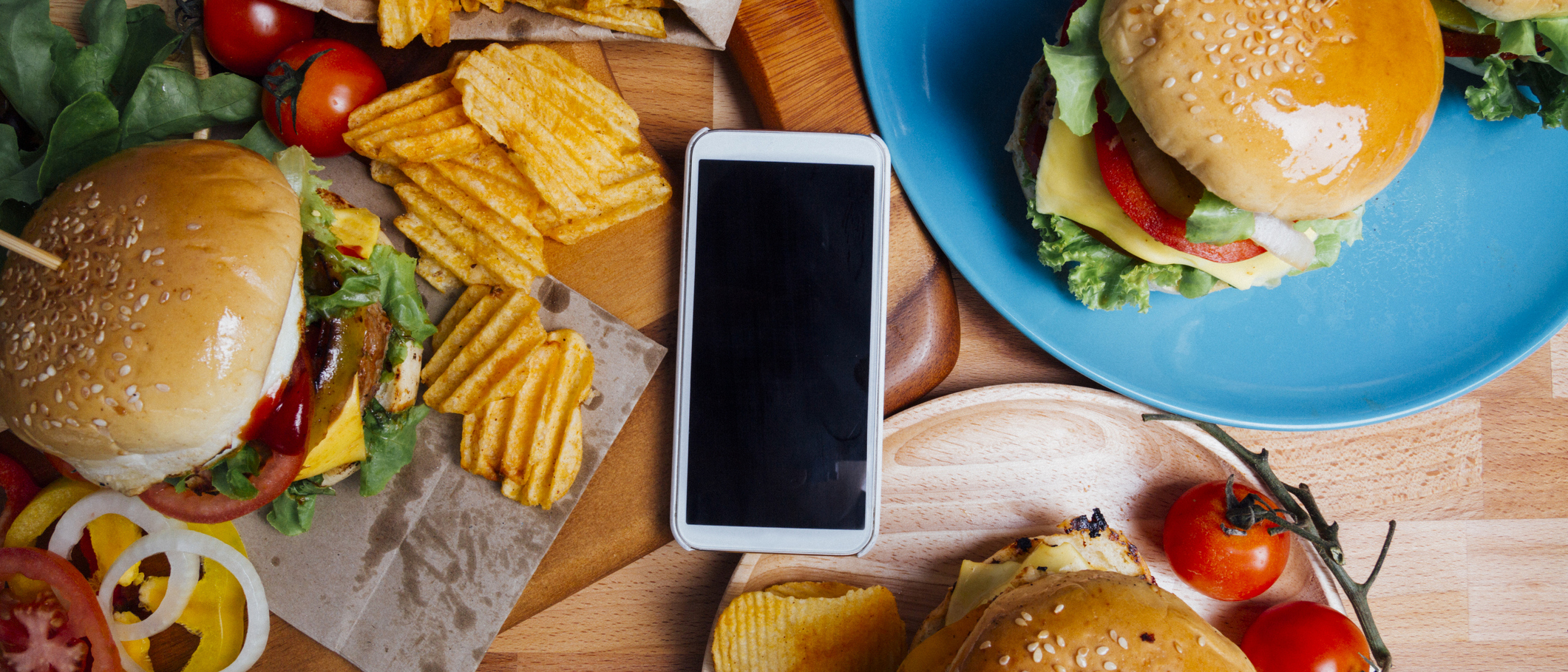 Cell phone placed in middle of a restaurant table surrounded by burgers and fries.