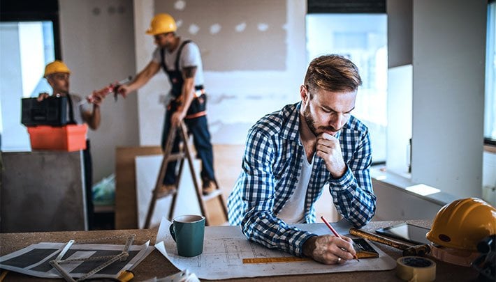 Male contractor drawing blueprints with co-workers working in the background